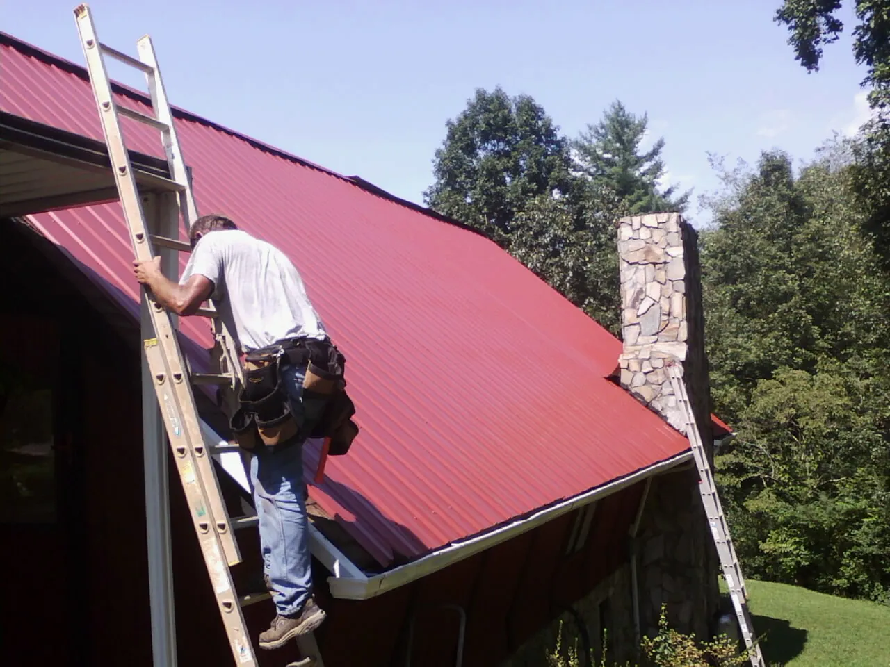 Roofer on a ladder beside a red metal roof and stone chimney — Soffit & Fascia Repair by Roofing Brothers in Moyie Springs